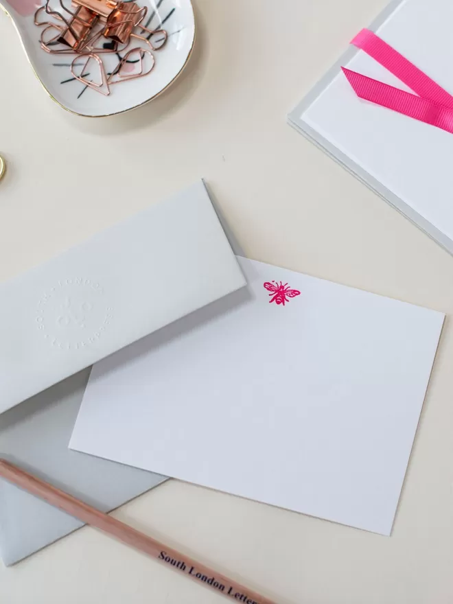 South London Letterpress Neon Pink Bee Notecard seen with a grey envelope and stationary on the desk.