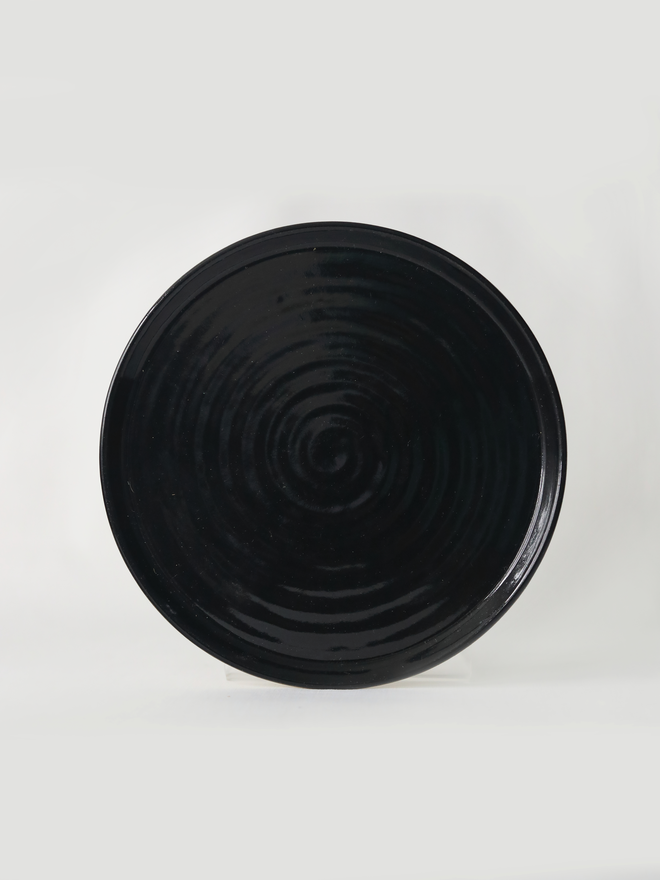 A handmade ceramic side plate in jet black glaze, photographed from the front against a plain white background, showing a natural concentric swirl texture from the wheel-throwing process