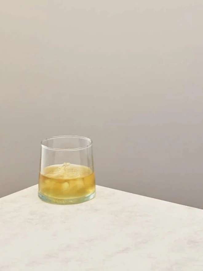 Low Glasses (Set Of 1 Or 4), a low glass sitting on a white counter top against a plain backdrop. It is filled by a soft drink. 
