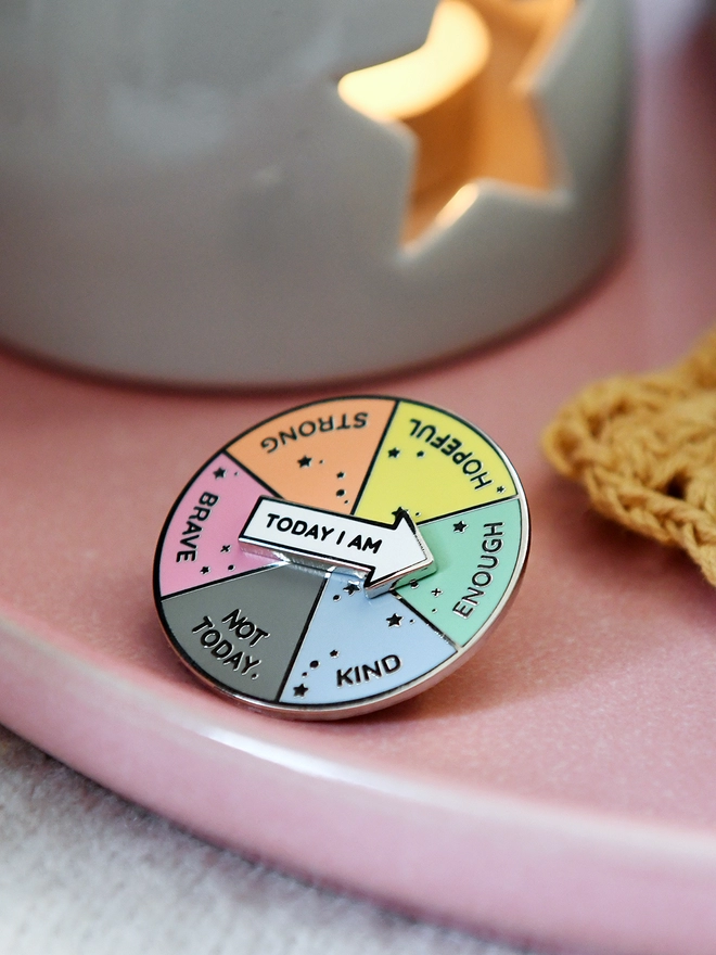 A round enamel pin that looks like a pie chart with six segments, each with positive word, and a white arrow that reads "Today I Am", is resting on a pink plate beside a candle.