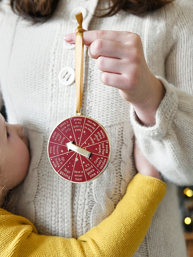 A girl is holding a deep red and gold Christmas decoration with a young child looking on. It has 12 segments, each one with a different Christmas activity idea, and a golden arrow in the centre.