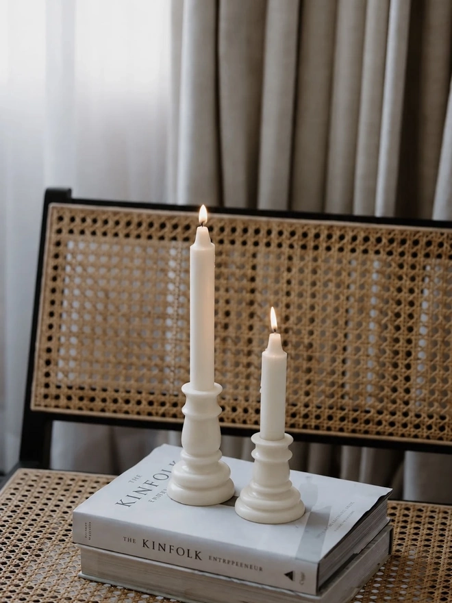 Baluster Christmas Candles (Set Of 2). Two cream candles moulded in the shape of classic candlesticks, standing on a stack of books on a woven cane chair against linen curtains. The all-in-one design looks like a candle in its holder, so no separate stand is needed.