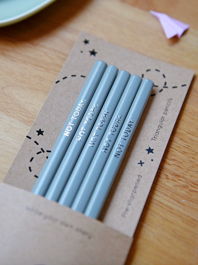 Five grey pencils with the words Not Today written along the side, sit within cardboard packaging on a wooden desk with stationery items.