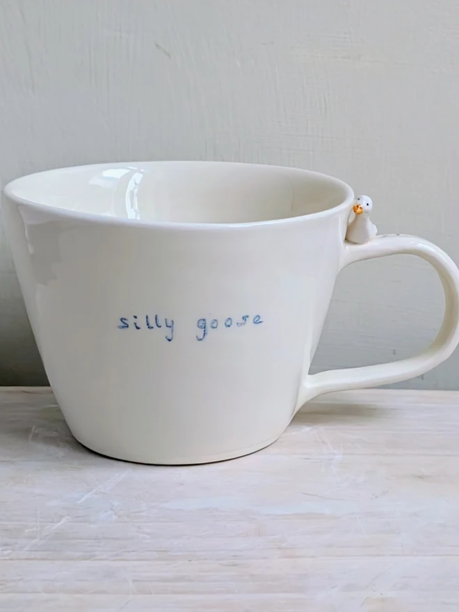 white handmade ceramic goose cup with a tiny bird, bird prints and the words silly goose handpainted in blue simple font