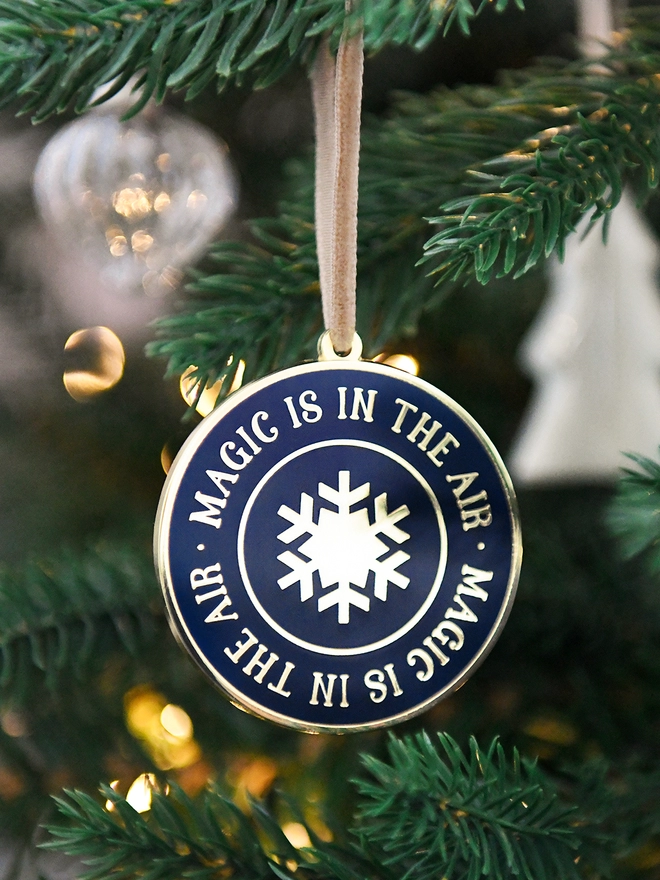 A navy blue and gold enamel Christmas decoration, with the words “Magic Is In The Air” surrounding a gold snowflake, is hanging on a Christmas tree.