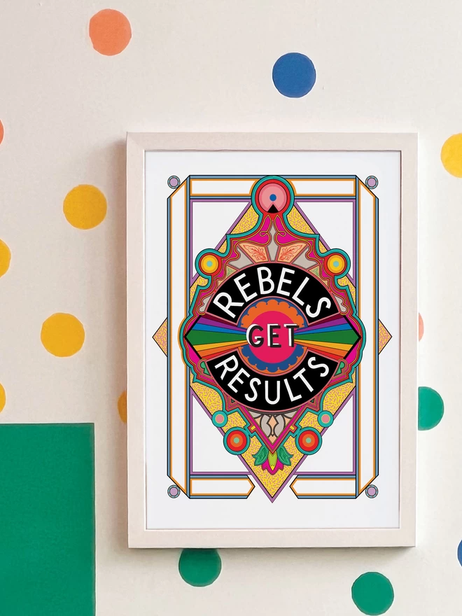 Rebels Get Results is written in white on a black background at the centre of this vibrant, abstract portrait illustration, with a white background and rainbows emitting from the centre, and multi-coloured detailing. The picture is hanging in a white frame on a white wall, with yellow, orange, green and blue spots and a green and yellow rectangle painted in the top left hand corner.