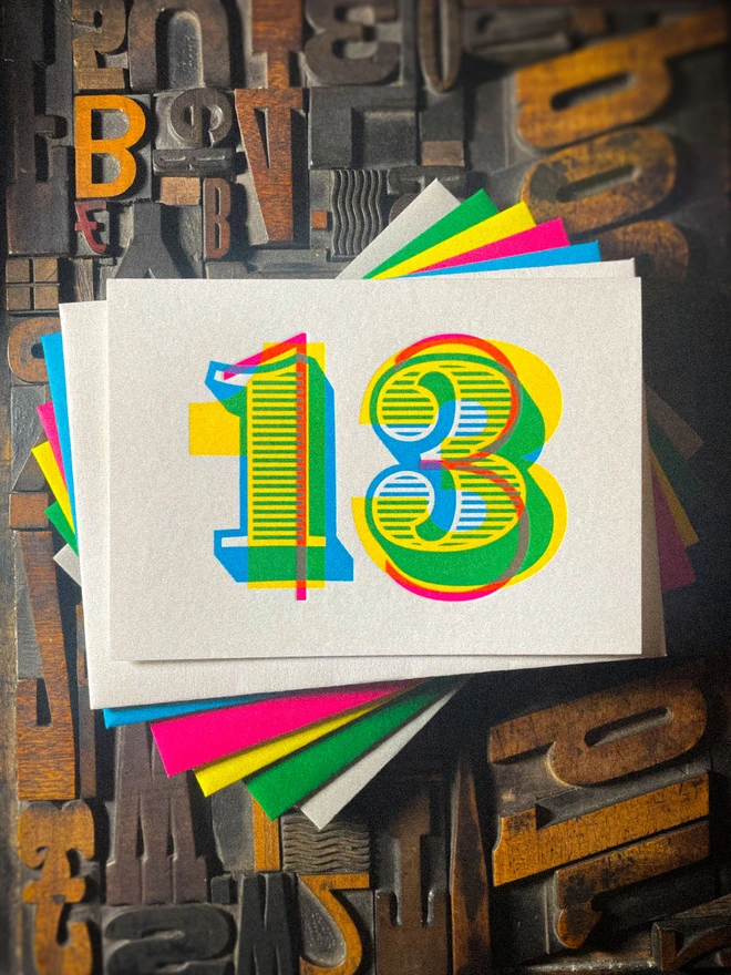 Handmade 13th birthday letterpress card featuring a bold number 13 printed in overlapping cyan, magenta and yellow inks, creating vibrant secondary colours. Printed on luxury GF Smith Colorplan Pristine White card and shown with colourful envelope choices against vintage wooden letterpress type.