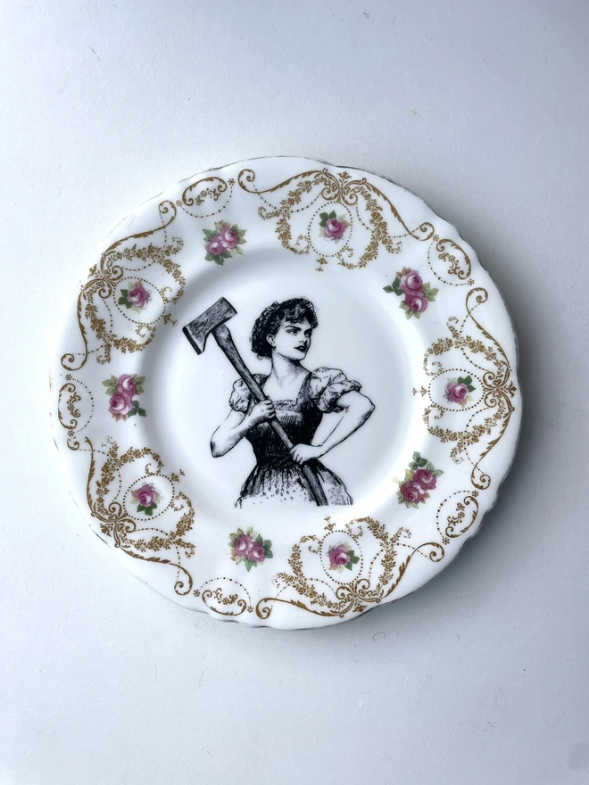 a vintage plate with a really ornate pretty border has a vintage black and white illustration of a beautiful girl in peasant clothes wielding an axe