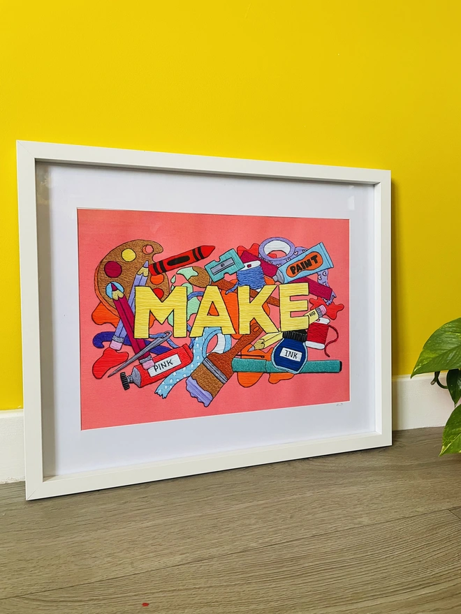 Colourful print of hand embroidered art reads MAKE surrounded by art supplies