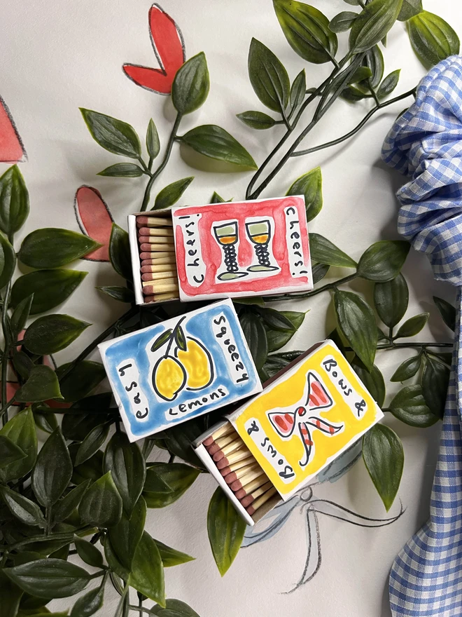 A collection of three hand-painted matchboxes with playful and colorful designs: one features two glasses with the word "Cheers!" written in bold lettering, another displays bright yellow lemons with "Easy Squeezy" written alongside, and the third showcases a red bow with the phrase "Bows & Arrows." Each matchbox is crafted with vibrant illustrations and creative lettering.