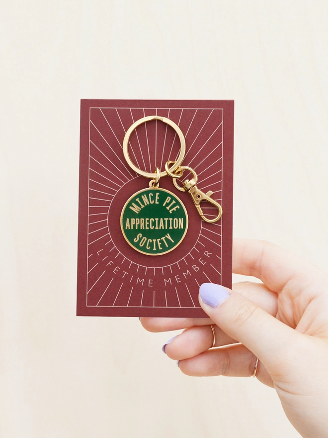 Hand holding out a Green enamel keyring featuring 'Mince Pie Appreciation Society' design