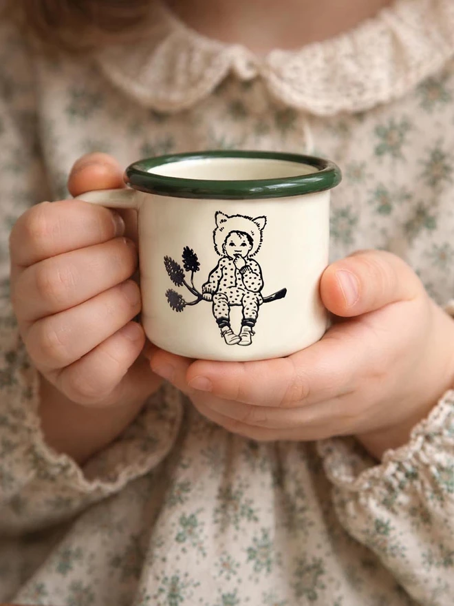 a small child holding a miniature enamel mug cream in colour with a bottle green rim featuring an illustration of a small cute pixie child sat on a pine cone branch wearing a wolf hat