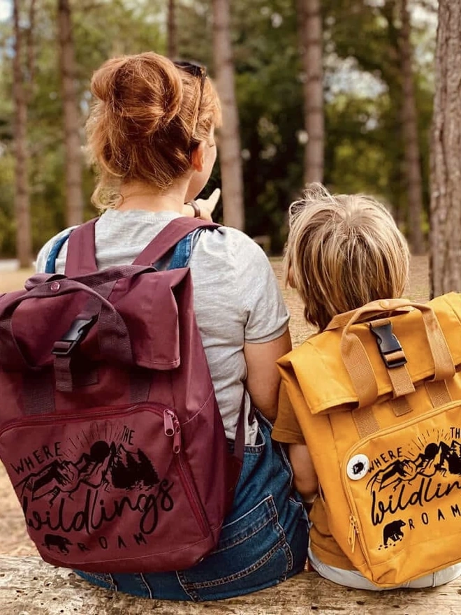 'Wildings' Children’s Recycled Roll Top Back Pack, two children sitting in a forest with colourful backpacks. 