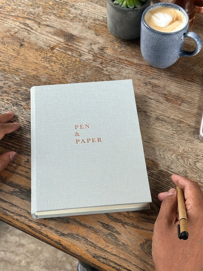 on a wooden table there is a pale grey linen notebook. The notebook has been printed with the words PEN to PAPER on the front. There is a cup of coffee in the background.
