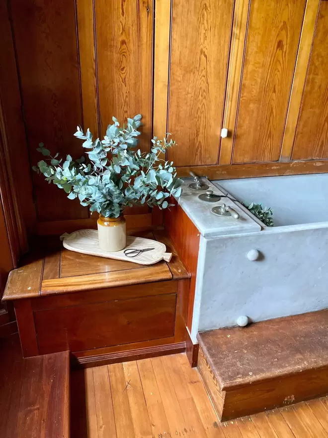Freshly Cut Eucalyptus Populus Greenery sits in a vintage brown ceramic vase. The vase sits on a light oak board with a pair of small vintage scissors alongside. The scene is set in a wooden panelled bathroom with a grey marbled bathtub in the background.