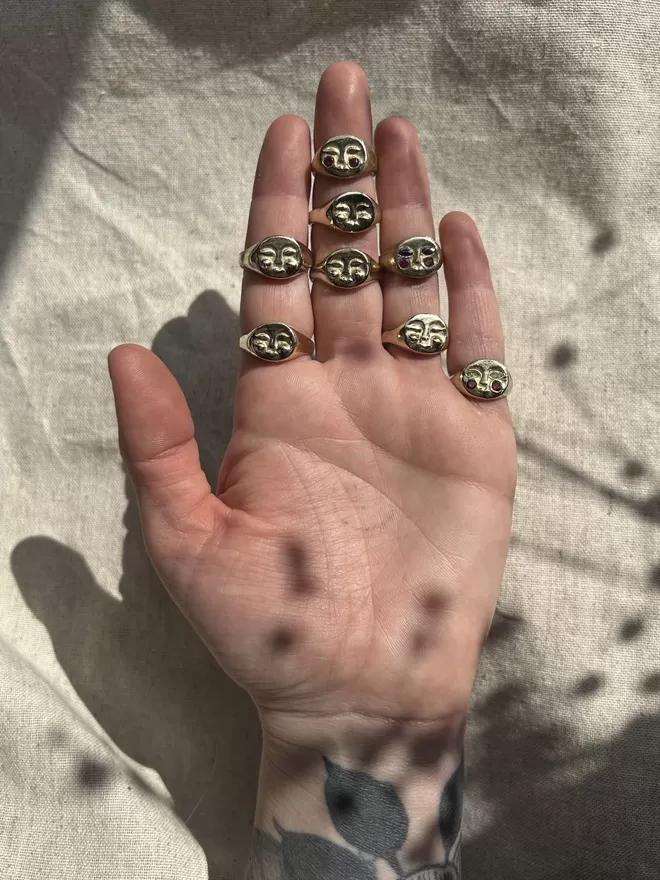 Image of a hand held up with the palm facing towards the camera. 8 rings are being worn on the fingers of the hand, all rings are gold coloured and some with precious stones for eyes and cheeks. The background is a cream linen fabric and there is a shadow of a plant overlaying the image 
