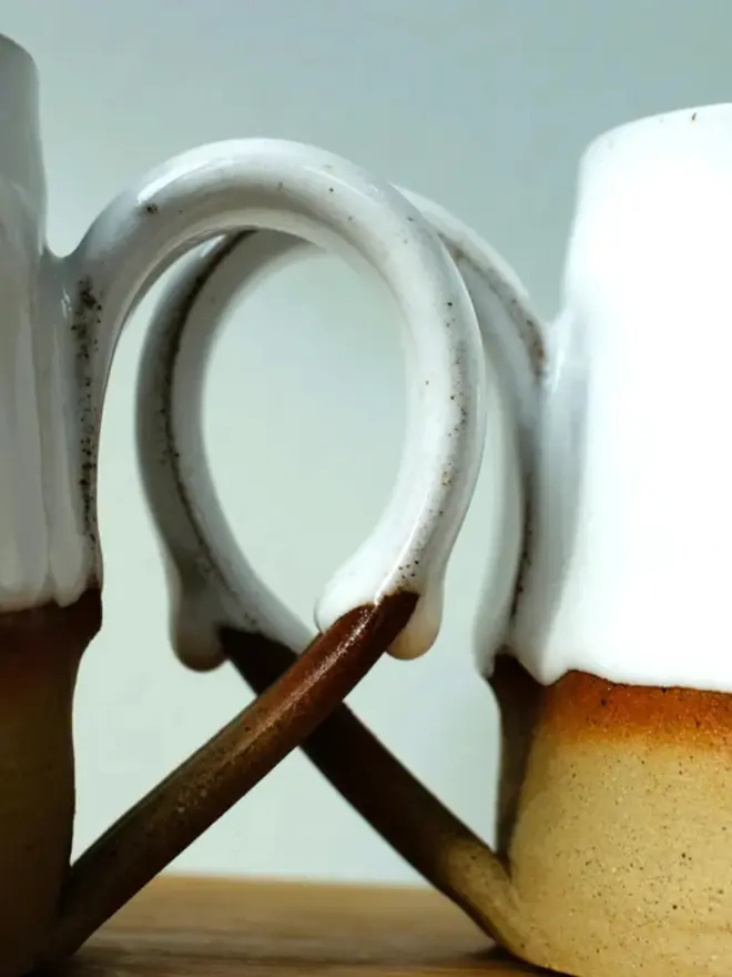 Handmade ceramic mug with a glossy white upper glaze and a natural toasted clay base, against a soft green wall background.