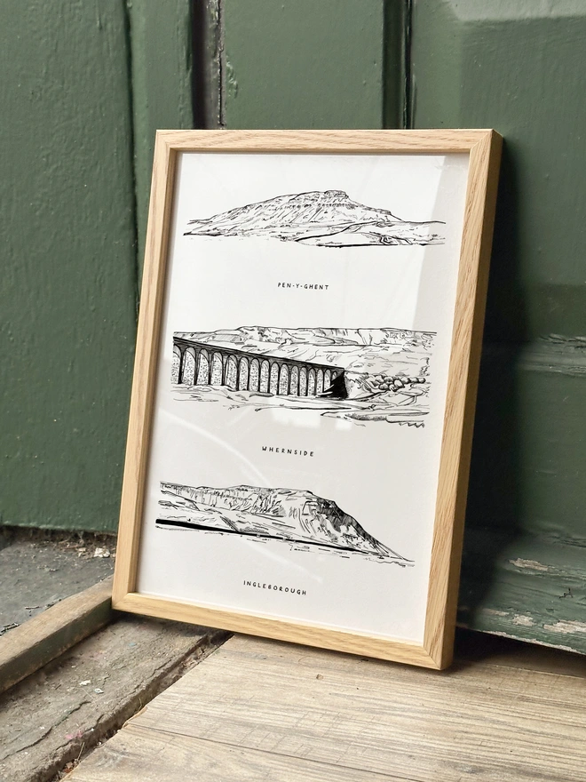 print featuring black and white line illustrations of the yorkshire three peaks, framed and leaning against a green door
