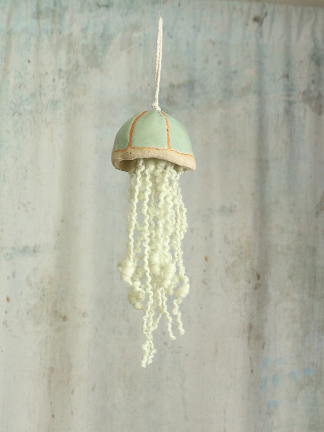 mint green handmade jellyfish hanging decoraton with green curly wool tenticles is handing in front of a mottled background 
