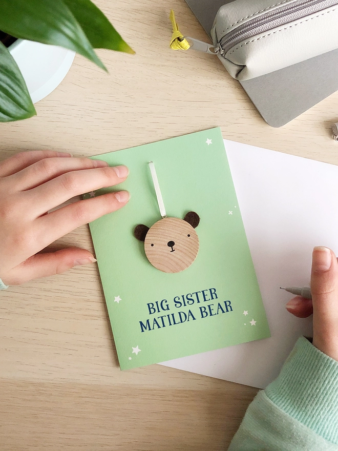 A green greetings card with a small wooden bear keepsake and the words "Big sister Matilda bear" printed on is on a wooden desk. 