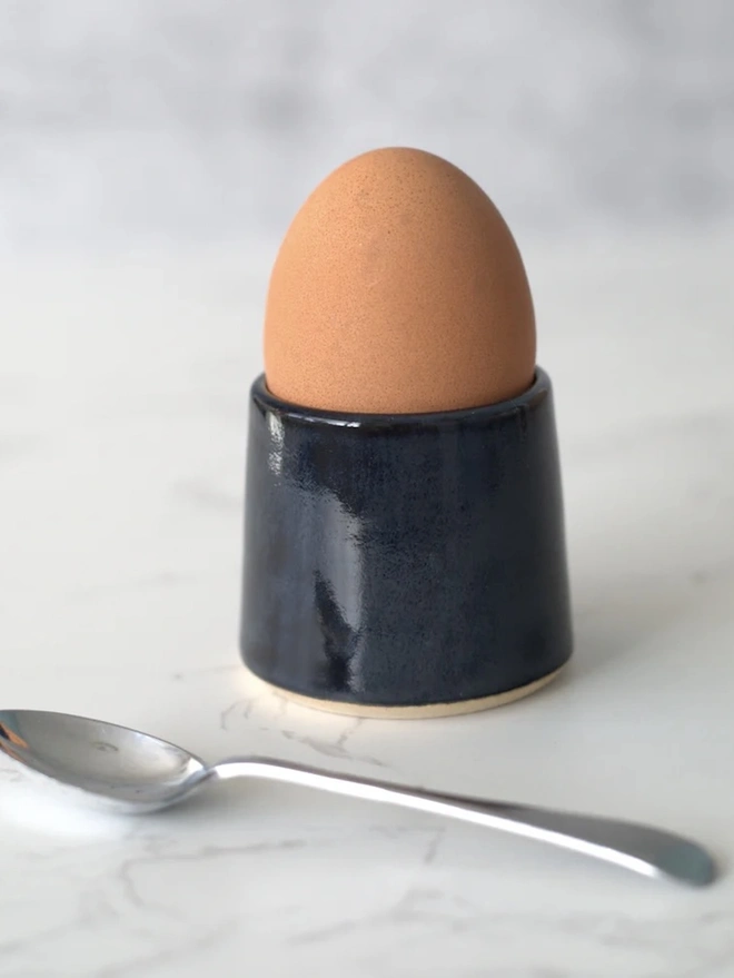 Classic Handmade Egg Cup, a ceramic egg cup sitting on a plain surface against a grey backdrop. It is accompanied by a spoon. 