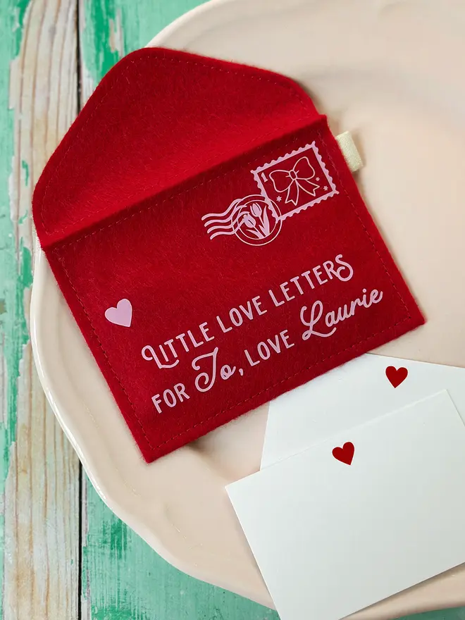 A handmade personalised red felt love letters envelope displayed on a vintage-style plate with small heart cards, styled on a rustic green wooden table for Valentine’s Day.
