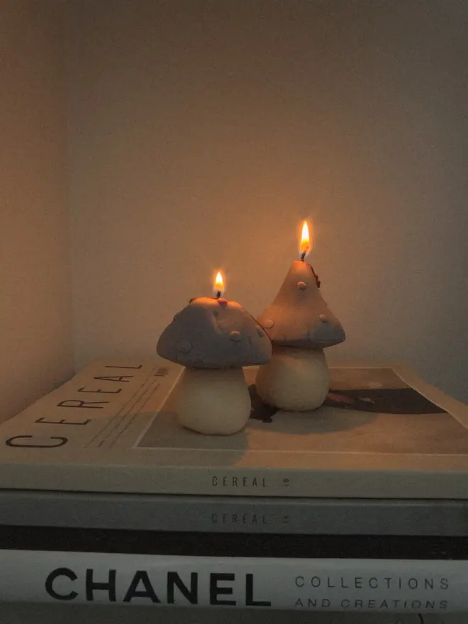 Pair Of Little Mushroom Candles (Set Of 2). Two lit mushroom candles with dotted caps glowing softly on a stack of coffee table books. The warm candlelight shows off their sculpted shapes, ideal for creating a calm, cosy atmosphere.