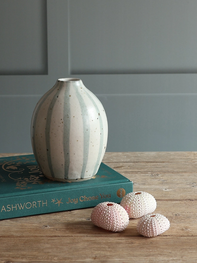 Handmade Ceramic Stoneware Vase in Blue and White Stripe. The vase sits on a teal hardback book with a collection of three pink sea urchins sat beside it on a wooden surface. 