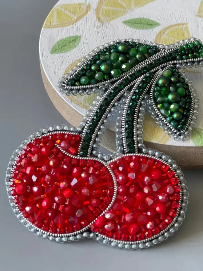 Red Cherry Beaded Brooch displayed on a grey table next to a dish with painted lemons. 