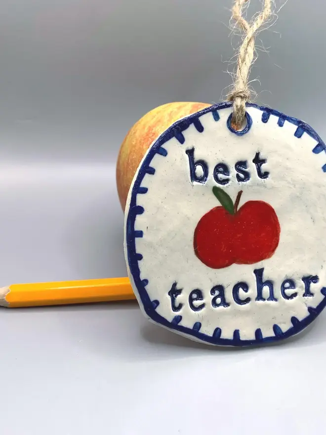 'Best Teacher' Handcrafted Ceramic Ornament, a ceramic apple ornament sitting on a plain white surface joined by an apple. 