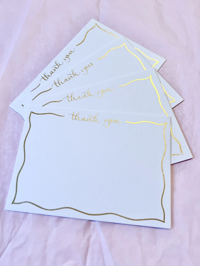 Four white notecards with thank you written in gold calligraphy at the top, and a gold wavy border around the edge of the card.