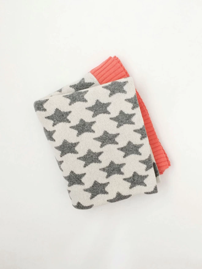 A folded junior blanket, photographed from above, showing a grey and white star pattern and a glimpse of coral pink trim.