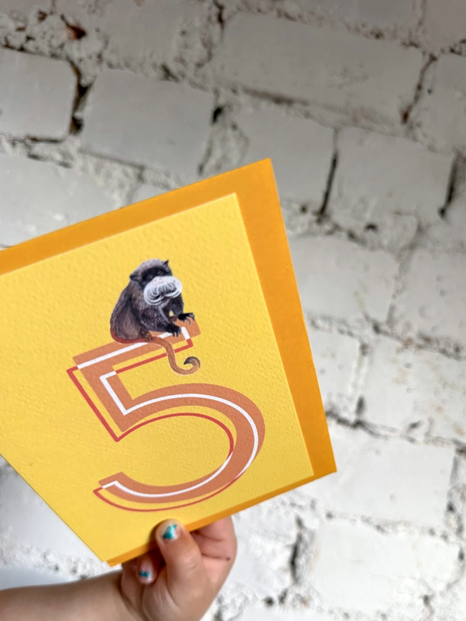 bright yellow card featuring a big orange number 5 and an illustration of an emperor tamarin with a bright yellow envelope