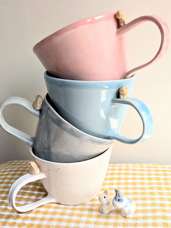 a stack of pink blue grey and white bunny cups with a tiny beige bunny on each handle and tiny pawprints on a yellow gingham cloth