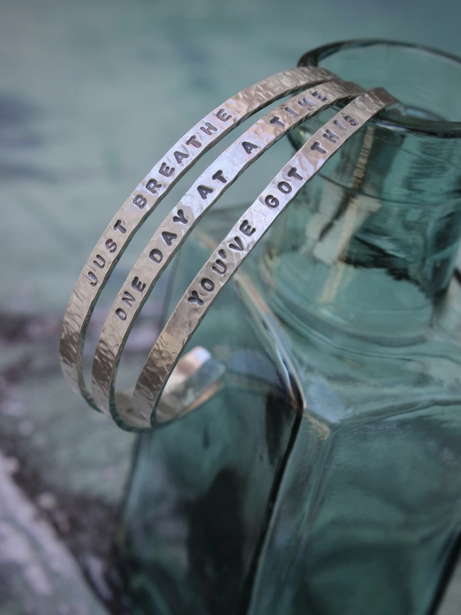 Three sterling silver cuff bangles with stamped text, balanced on the neck of an antique green teal bottle, against a green background.