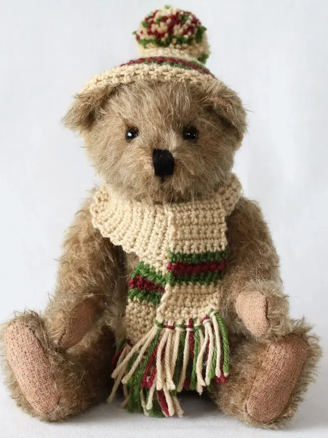 Brown teddy bear sitting upright against a white background, wearing a knitted beige hat and scarf with green and burgundy details.