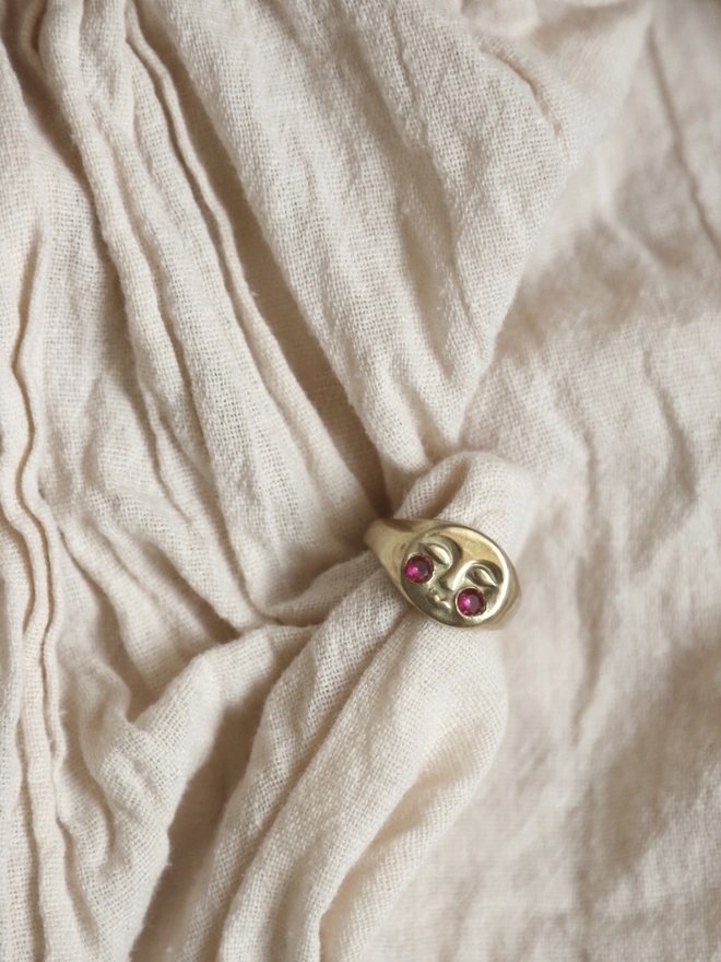 Image of a gold moon face ring with ruby encrusted cheeks, laying on a cream linen type fabric 