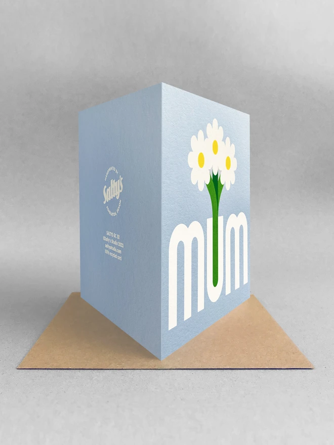 Rear view of a light blue square card with mum printed in grey, the U is a vase with three flowers in it. Card is stood on a brown Kraft envelope, all in a light grey space with soft grey shadows.