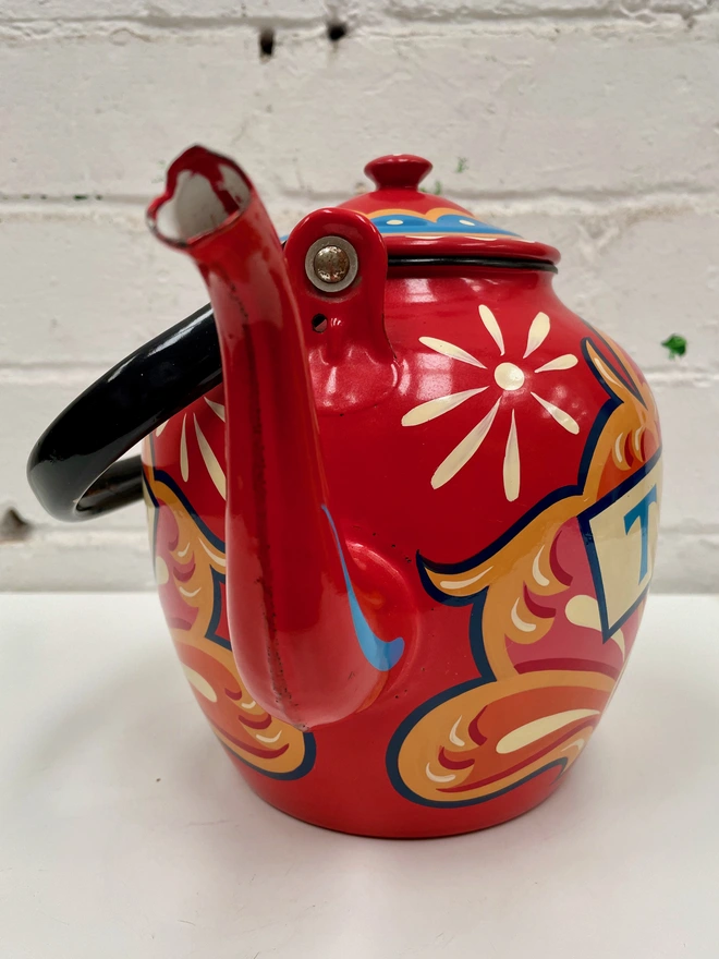 Red vintage enamel teapot with folding pouring handle, hand-painted with a traditional fairground inspired design in pink, orange and yellow. 'Tea' is painted in a banner across the centre.