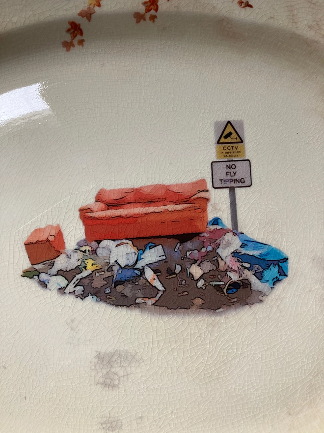 large oval vintage plate featuring a cottage on the left with a floral border. In the centre sits a pile of rubbish with an old sofa on top, next to a sign stating 'no fly tipping'.