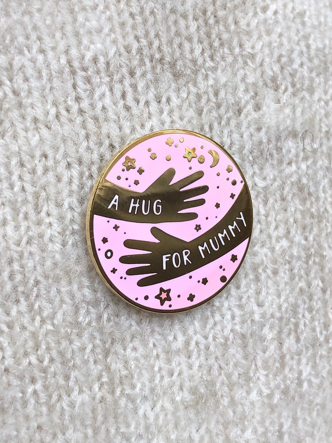 An enamel pin badge that has a hugging arms design and the words "A hug for Mummy" is pinned to a beige jumper.