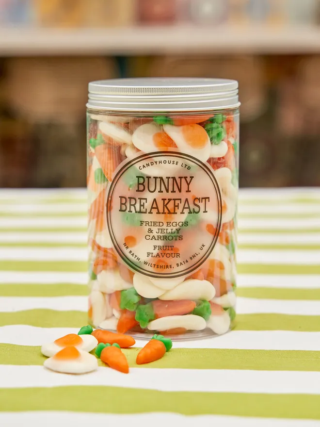 Extra large jar of fried egg sweets and jelly carrots, with a silver lid on a green and white striped tablecloth in an outdoor setting