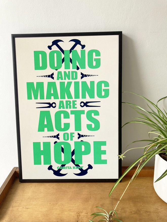 Framed green and blue typographic print. The Corita Kent quote reads "Doing and making are acts f hope." Navy blue tools are placed around the green text. The print rests on a shelf next to a spider plant.