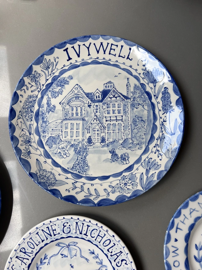 Blue and white hand-painted place with house portrait and "Ivy Well" at the top with a botanical border