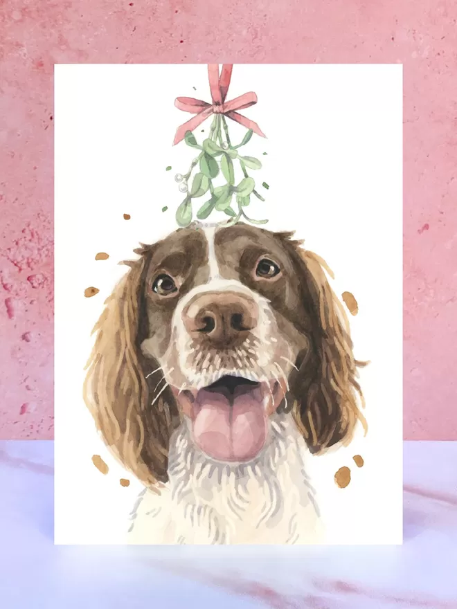 A Christmas card featuring a hand painted design of a Springer Spaniel, stood upright on a marble surface.