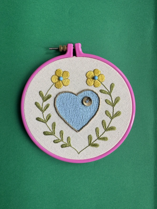 A pink embroidery hoop containing a hand stitched blue heart surrounded by two yellow flowers on long green stalks