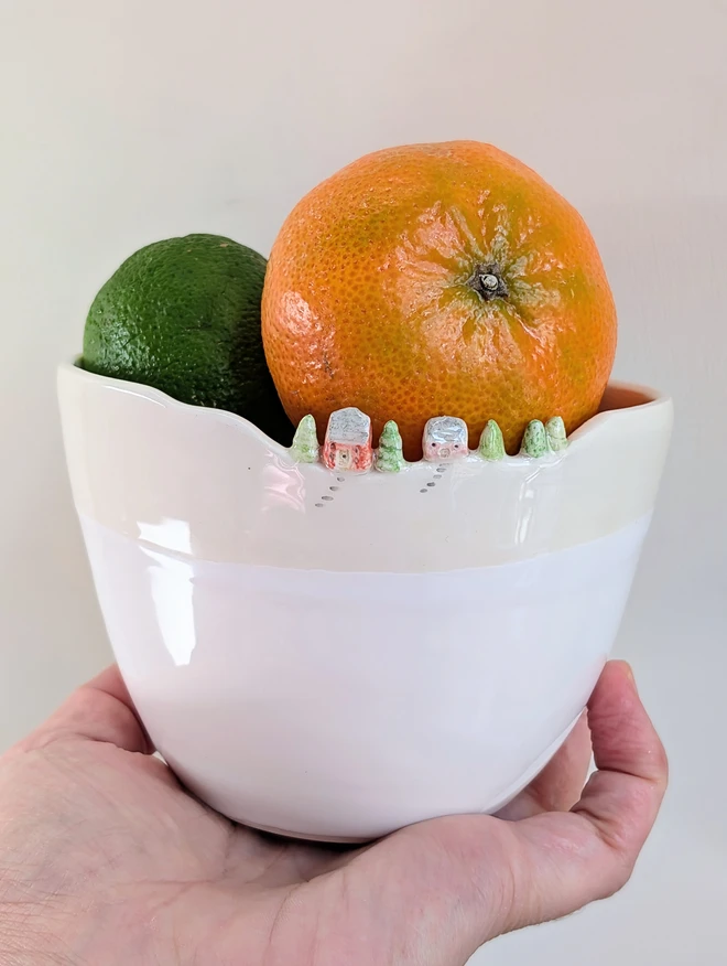 Handmade small ceramic white bowl with a tiny pink and red house and tree details on the rim and white glaze held aloft in a hand with a fruit inside 