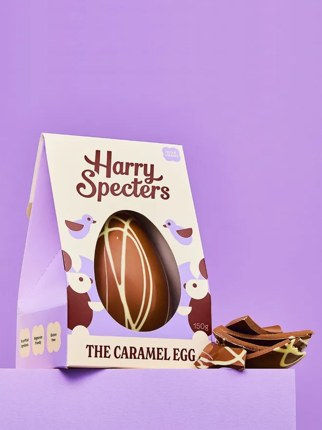 A caramel chocolate Easter egg on a purple background, next to broken caramel chocolate Easter egg pieces