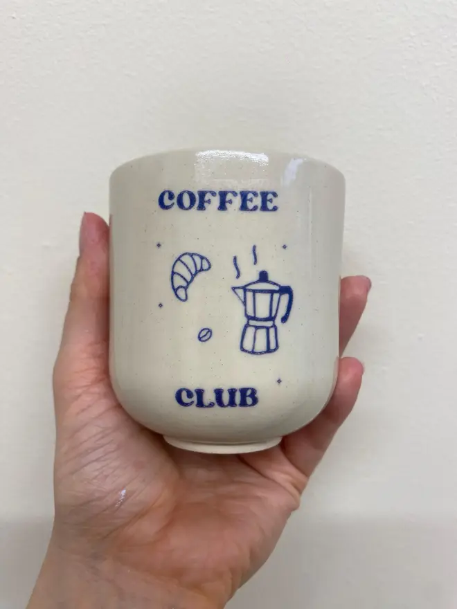 'Coffee Club' Handmade Ceramic Coffee Cup, a coffee cup held against a white backdrop with a blue design. 