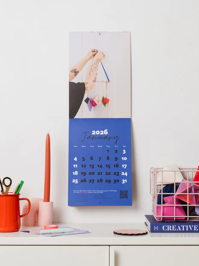 Calendar hung on wall with cabinet below, with a jug, candle, book & basket styled around it.